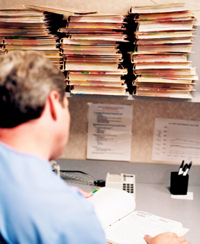 A male nurse sitting at a desk with a lot of folders above him on a shelf